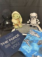 Photo showing three Star Wars themed stuffed animal pillows (Darth Vader, Yoda, Stormtrooper), a Star Wars blanket, a Pottery Barn Kids pillow with the phrase 'May the Force Be With You', and two Star Wars print sheets.