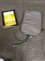 Amazon Kindle eReader, black, showing startup screen with 'Please verify Kindle is registered' prompt. Next to it is a black padded sleeve with some wear marks and black USB charging cable.