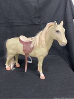 Full side view of the beige plastic toy horse with cream-colored mane and molded reddish brown saddle missing one stirrup, standing against a black fabric background.