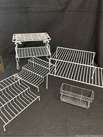 Photo of five wire shelf organizers including three white tiered stackable shelves, one large white wire shelf, and one small chrome utensil organizer basket arranged against black backdrop.
