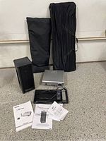 Photo showing the projector, speaker, Blu-ray player, remote controls, cables, and instruction manuals arranged on a garage floor, along with two black bags likely for the screen or stands.