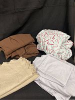 Four distinct bedding pieces including a brown knit sheet set, a candy cane flannel sheet set in white with red candy cane prints, a grey and white striped/checked moleskin sheet set, and a cream-colored scalloped matelasse quilt.