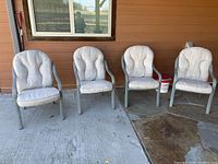 Four lightweight painted metal outdoor chairs with light gray cushions lined up on a concrete patio surface next to a brown wall.