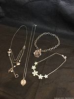 Five silver toned necklaces and one bracelet arranged on black fabric, showing overall design and variety.
