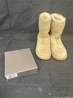 Ugg Classic Short II boots in buff color shown with original sheepskin care kit box on black fabric background.