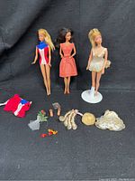 Three vintage Barbie dolls and assorted accessories on black backdrop including Gold Medal Barbie, Brunette Barbie, and Ballerina Barbie with outfit pieces and shoes.
