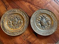 Two smaller brass plates featuring repousse fruit designs including grapes, pears, and apples, with decorative borders.