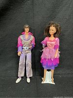Two dolls representing Donny and Marie Osmond, dressed in original colorful outfits with microphones, one in a two-tone purple outfit and one in a multi-layer pink and purple dress standing on a white stand