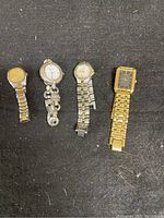 Four wristwatches laid out on dark surface; three ladies watches and one mens watch; gold and silver tones; one with cracked crystal; all need batteries