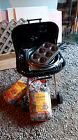 Black BBQ grill with lid open. Two fry pans and two muffin tins placed inside the grill. Two bags of charcoal briquettes in front of grill.