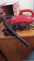 Red Dirt Devil Scorpion hand vacuum on wooden surface with attached hose and power cord coiled beside it.