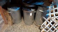 Photo of three aluminum garbage cans with lids and side handles stored on a garage floor with gravel and wooden walls.