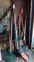 Full set of garden tools leaning against wall including shovels, rake, brooms, and other hand tools.
