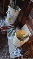 Photo showing two white plastic buckets on a wooden shelf, one bucket filled with black and metal lawn markers, the other bucket containing small gardening tools. Several gardening shears and pruning tools also placed on the shelf nearby.