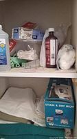 Shelf with medical and cleaning supplies including rubbing alcohol, cotton balls, cotton swabs, nasal spray, and a boxed steam and dry iron below folded linens.