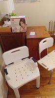 Two white plastic bath/shower chairs and boxed Silka Sonic Clean Plus face cleaner on wooden floor, near wooden furniture.