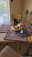 Dining table with four southwestern style placemats, wooden lazy susan holding napkin holder, salt and pepper shakers, cast iron candle holders with candles, and black metal leaf bowl.