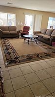 View of the beige area rug placed in a living room between sofas and chairs, showing the geometric and leaf design border.