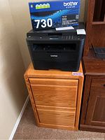 Photo showing Brother HL L2395DW black printer with Brother TN730 toner cartridge box on top of oak wood filing cabinet.