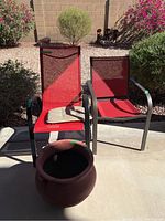 Two red mesh outdoor chairs with black metal frames on a concrete patio with brown clay pot in front. Background with plants and garden area.