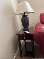 View of wood end table with ceramic lamp and beige shade on top next to a red sofa.