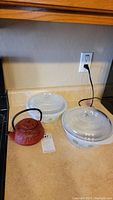 Two Glasbake glass casserole dishes with lids and a red metal teapot on kitchen counter