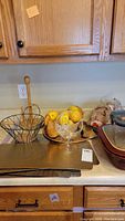 Countertop with ceramic baking dishes stacked on one side, wire and wicker basket, decorative stemmed glass dish with faux oranges, paper towel holder, kitchen towels, and counter protector