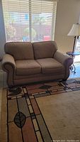 Full view of the beige cloth love seat placed near a window, showing the overall shape and size.