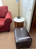 Wood side table with round marble top, small brown pleather ottoman, and black desk lamp in corner near red chair and door.