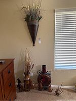 View of room corner with metal wall sconce, amber pitcher with faux flowers, and metal Howling Wolf sculpture on floor near wooden dresser and window.