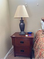 Nightstand with two drawers and ceramic lamp with beige lampshade on top, standing next to bed