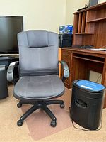 Grey cloth office chair in front of wooden desk, adjacent to black Royal paper shredder on carpeted floor.