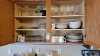 Kitchen cabinet open showing organized Mikasa Ocean Jewel White dinnerware plates and bowls on top shelves, with coffee mugs and short glasses on the bottom shelf. Clear stemmed wine glasses on top shelf.