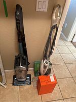 Two cleaning machines side by side on tile floor: Shark vacuum on left, Shark steamer on right, along with boxes and accessories.
