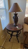 Side table with round wood top and clay lamp with shade placed on top beside window and couch.