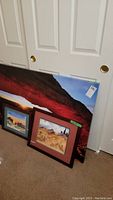 Three pieces of southwest inspired wall art leaning against a white door, including a large canvas print and two framed prints, all showing desert and rock landscapes.