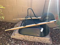 Side-angle photo showing wheelbarrow from above with black metal tray, two long wooden handles, black metal handle supports, and one black pneumatic tire.