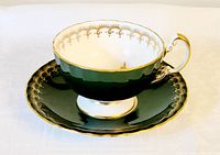 Tea cup and saucer set displayed on white fabric, showing dark green body with white and gold trim interior and exterior
