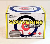Photo of the souvenir box with curling stone graphic and 'Souvenirs That Don't Suck' text, showing branding and product packaging.