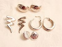 Four pairs of sterling silver earrings displayed on a cream fabric background: engraved half-hoop, abstract zig-zag, smooth rounded hoop, and ribbed dome-shaped studs.