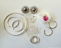 Overview of sterling silver jewelry lot showing decorative stud earrings, multiple pairs of silver hoop earrings, delicate chain necklace and pink heart pendant in a clear display case on white background.