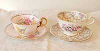 Two teacup and saucer sets displayed on fabric. The left Aynsley cup has pink and yellow floral designs with gold trim. The right Queen Anne cup has a more intricate floral garland pattern with pink bows and gold accents.