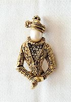 Front view of vintage costume jewelry brooch portraying a woman with textured hat, faux pearl face, rhinestone decorated jacket, and crossed hands.