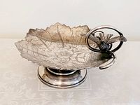 Front-left angle showing overall leaf shape, engraved floral design, hummingbird handle, and pedestal base.