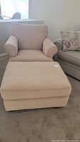 Photo of beige upholstered armchair and matching ottoman side by side on carpeted floor, highlighting fabric and shape.