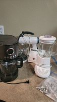 Photo showing black Cuisinart programmable coffee maker with glass carafe, white Cuisinart blender, and white Sunbeam hand mixer on floor, including coffee pot box behind blender.