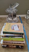 Stack of assorted cookbooks next to Cuisinart food processor parts on carpet.