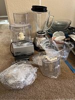 Cuisinart food processor with multiple attachments and Oster blender with additional smaller bowls and plastic covers, arranged on floor.
