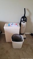 Photo of Dirt Devil vacuum, white laundry hamper, white rectangular wastebasket, and black mesh wastebasket grouped together on carpet.