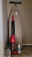Photo showing Dirt Devil vacuum, ironing board, Black and Decker iron, and hair dryer all together leaning against wall.